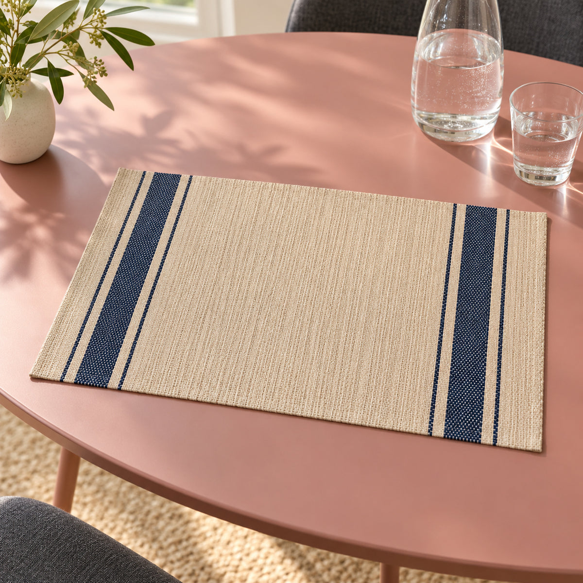 Beige rectangular table placemat with narrow and broad navy blue stripes down both ends, displayed on a muted pink dining table