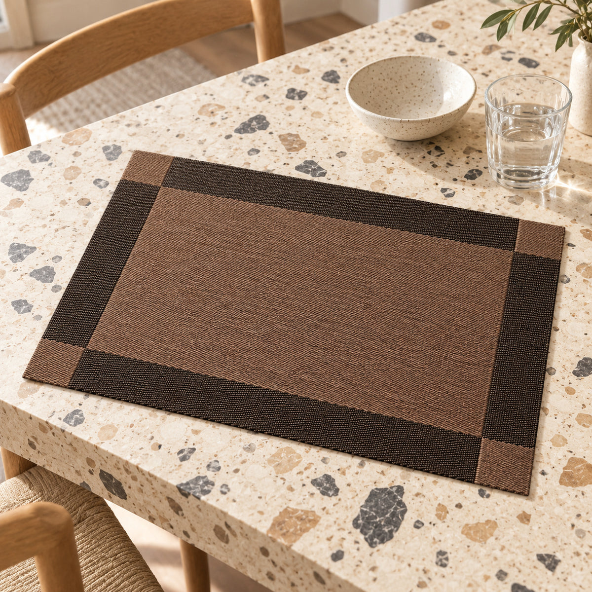 Cocoa brown rectangular placemat with wide charcoal black border and fine dark inner framing on a pale terrazzo dining table