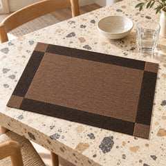Cocoa brown rectangular placemat with wide charcoal black border and fine dark inner framing on a pale terrazzo dining table