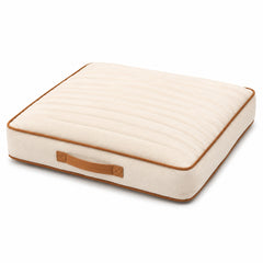 Ivory square channel-quilted floor cushion with tan piping and carry handle on white background