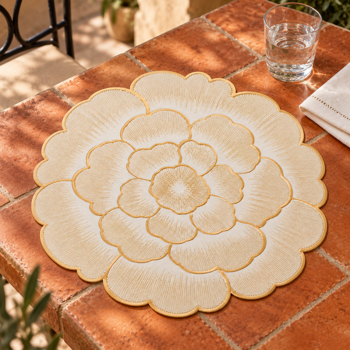 Ivory flower-shaped dining placemat with layered rounded scalloped petals and fine gold-coloured outlines on a terracotta tiled table