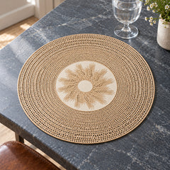 Round natural beige placemat with an open-weave-looking circular border and ivory floral sunburst medallion at the centre on dark marble