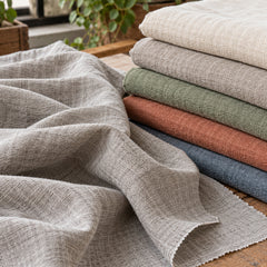 Soft washed grey-beige linen draped beside layered neutral and earthy fabric colours in a natural-light studio; Primeval sourcing reference PML-FAB-007