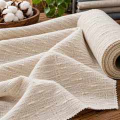 Natural cream cotton slub fabric spread in textured folds beside a fabric roll, showing irregular raised yarn detail; Primeval fabric reference PML-FAB-008
